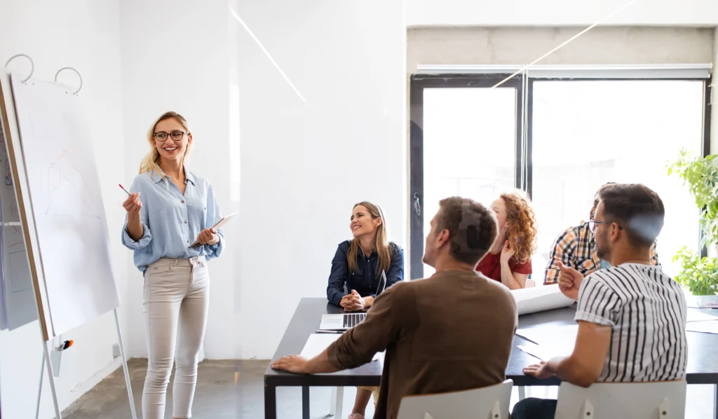 Teammeeting mit 6 Personen - eine Frau mit Brille steht an einem Flipchart und zeigt etwas, während die anderen interessiert zu ihr blicken.